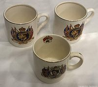 Three ceramic coffee mugs commemorating British royal coronations, two with King George VI and Queen Elizabeth 1937 coronation portraits and one with Queen Elizabeth II, positioned on white surface.