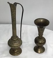 Two antique brass vases side by side, taller with handle on left, shorter with black outlined engravings on right.