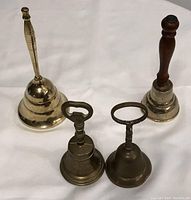 Four brass hand bells arranged on white surface, showing the varying height and handle styles clearly.