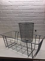 Two wire baskets shown together against a light brick wall, one large rectangular basket and one smaller cylindrical waste basket inside the larger one.