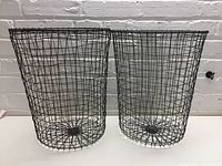 Two black metal wire baskets side by side on a white surface against a white brick wall background.