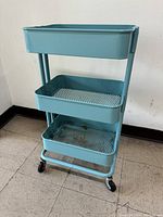 Three-tiered light blue Ikea rolling cart standing on tiled floor.