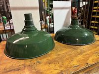 Two green metal spinning enamel industrial light shades side by side on a wooden surface showing differences in size and top mounts.