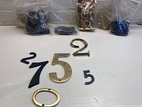 Photo showing several loose house numbers on a surface including brass and black plastic numbers, along with 6 plastic bags containing more numbers in the background.