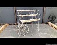 White three-shelf metal plant stand with ornate scroll and wheel designs, photographed against a gray backdrop.
