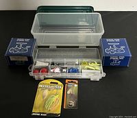 Full fishing lot showing plastic tackle box with compartments filled with assorted tackle, two boxed limited spool spinning reels, and two packaged spinnerbaits.