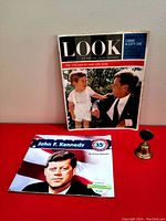 Photo showing the 1963 LOOK magazine with President John F Kennedy and son John Jr. on cover, the new children's book about JFK, and the vintage brass flag bell.