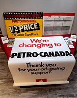 Photo showing multiple advertising signs including photofinishing half price sign, transition product chart, large 'We're changing to Petro Canada' sign, Fina Quebec maps and credit card applications on table