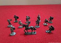Twelve assorted 1977 Ral Partha Dungeons & Dragons metal miniatures arrayed on red surface, each about 1 inch tall. Mix of humanoid fighters and one mounted figure.