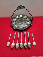 Seven small dessert spoons with various markings displayed in front of the bride's basket dish, showing their size and condition with tarnish.