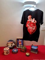 Full lot displayed including 7 assorted Christmas and Coca Cola collectible tins and 1 black T-shirt with Santa Claus Coca Cola graphic hung on wall