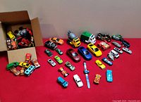 Large assortment of toy vehicles and wristwatch displayed on red surface, including various small cars, trucks, motorbikes, and a digital race car themed watch.