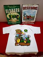 Photo of both metal signs and the John Deere t-shirt laid out on red surface.