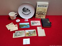 Wide view of entire lot on red background including book, plate, mugs, postcards, and diary.