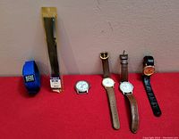 Five vintage men's watches displayed on red surface: one blue fabric band watch with REM TAI branding, one Timex watch in original sealed packaging, three leather strapped watches with white or orange dials, one with no band.