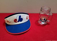 Photo showing the vintage Montreal Expos baseball visor and the glass mug side by side on a table, highlighting the visor's front panel and logo pins.