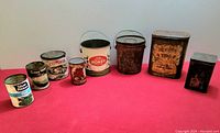 Full view of all 8 assorted vintage kitchen and pantry tins arranged in one row on a red surface with a white wall backdrop. Tins vary in size and shape from small circular cans to larger pails and rectangular tins. Visible text includes 'Golden Harvest', 'Sealtest Cottage Cheese', 'Farm Fresh Peas', 'Pure Honey', 'Salmon', and 'Tea'.
