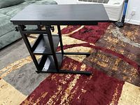 Black height adjustable snack table with two shelves and metal frame positioned on a carpet beside a couch, showing tabletop and storage shelves.