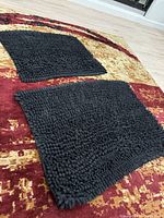 Two thick, textured black chenille loop bathroom rugs shown on a multi-colored floor mat, with visible non-slip backing texture and pliability.