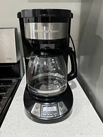 Front view of Hamilton Beach mid-size black and silver coffee maker on kitchen counter showing glass carafe, control buttons and digital display on base.