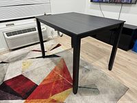 Photo showing the full angled view of the black dining table on a colorful geometric rug with light wooden floor in the background.