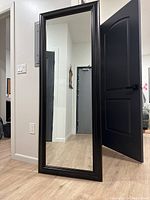 Full length mirror with bronze frame standing against a wall indoors, showing wood flooring and surrounding door frames.