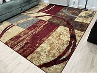 A new polypropylene area rug placed on light wood flooring in a room with a sofa, showing the rug's abstract geometric pattern in brown, gray, red, and beige tones.