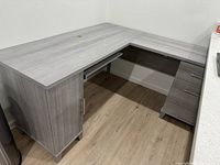 Photo showing full L-shaped workspace desk corner with pull-out keyboard tray and storage cabinet under left side, gray wood finish with metal handles.