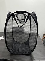 Front view of a black mesh foldable mini laundry hamper sat on a table against a plain off-white wall with a visible rectangular background object.