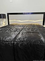 Full front view of the modern queen-size bedframe with black padded headboard, built-in LED light strip lighting up with a zig-zag pattern and shiny black comforter on the mattress.