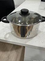 Silver-colored stainless steel pot with two black handles and clear glass lid, placed on white countertop.