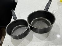 Photo of two black cooking pots with long handles shown on a white countertop, one smaller and one larger.