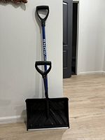 Full view of the snow shovel standing upright against a wall on light wood flooring, showing black blade, blue shaft, and black handles.