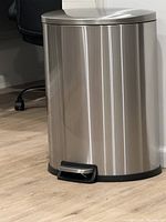 Silver oval-shaped trash can with foot pedal, placed on wooden floor near a wall and office chair.