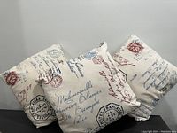 Three cream-colored sofa pillows with vintage script and postmark design leaning against a wall on the floor.