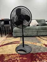 Photo showing a black standing pedestal fan positioned on a carpeted floor in a living room environment with furniture in the background.