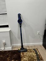 Blue cordless stick vacuum cleaner standing upright against a white wall, showing overall compact design and motorized brush head on floor.