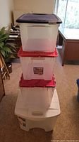 Stacked four plastic storage totes with lids in a room. Visible red lids and one blue lid on top. Good overall condition visible.