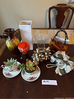 Full view of assorted decor items on a table including vases, faux succulent teacups, glass bowl, floral arrangements and ceramic purse vase.