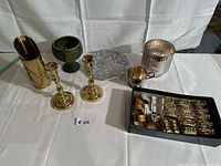 Lot of assorted decor on white cloth, including candlesticks, goblet, candle holders, chalice cup, napkin holders and brass container.