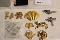 View of full collection of gold-tone and silver-tone brooches and pins on beige background including dog, flowers, basket, bird, bow, shell and rectangular rhinestone brooches.