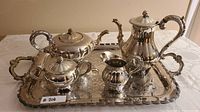 Photo of entire large silver tea and coffee set including teapot, coffee pot, sugar bowl, creamer, and a large silver tray with intricate engraving on white tablecloth.