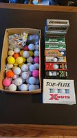 Photo showing a box of assorted loose golf balls in various colors next to stacked boxes of golf balls from various brands.