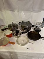 Photo showing the complete set of assorted pots and pans including stainless steel pots, cast iron skillet, ceramic pans in orange and beige, and glass cookware items.