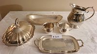 Full view of the silver water jug, cream and sugar bowls, covered bowl, and one of the two trays displayed on a table with cloth.