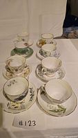 Overview photo showing all 8 tea cups and 8 saucers arranged in two rows on a white surface with neutral backdrop, showcasing the variety of floral and scenic designs and matching pairs.