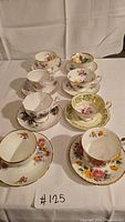 Seven fine china tea cups and saucers arranged on white cloth showing various floral designs and gold trim.