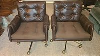 Front view of two brown leather office chairs with button tufting, padded armrests, and brass caster wheels.