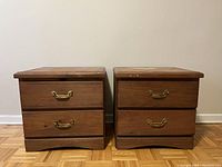 Front view of two wooden bedside tables showing two drawers each with brass-style metal handles and wood finish with surface wear.