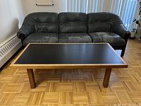 Photo of rectangular coffee table with black melamine top placed in front of a black leather sofa on a parquet floor.